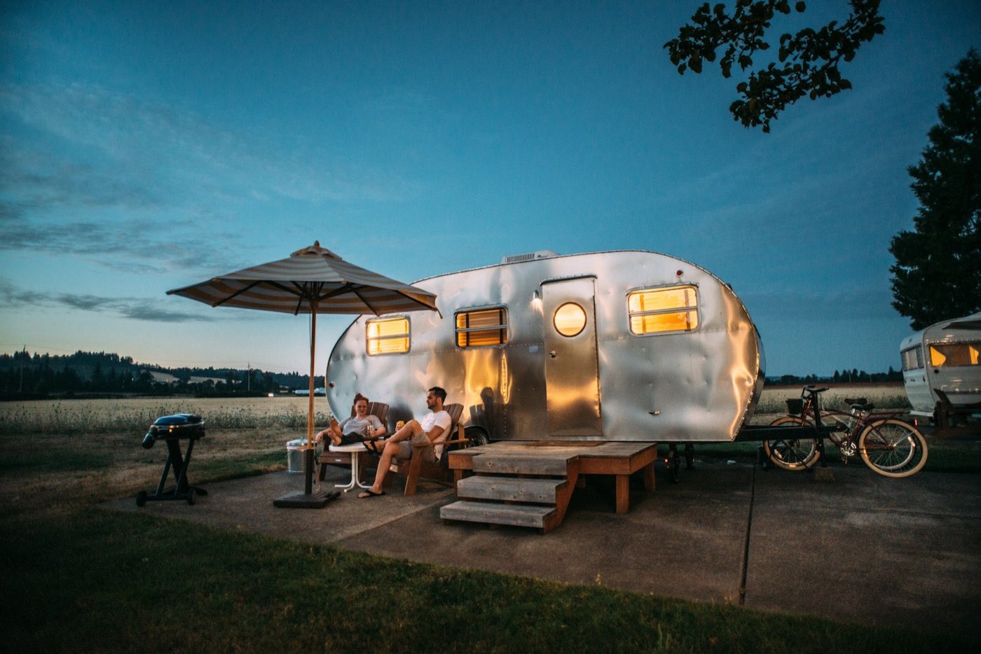 Travel trailer glowing at dusk with camp chairs outside